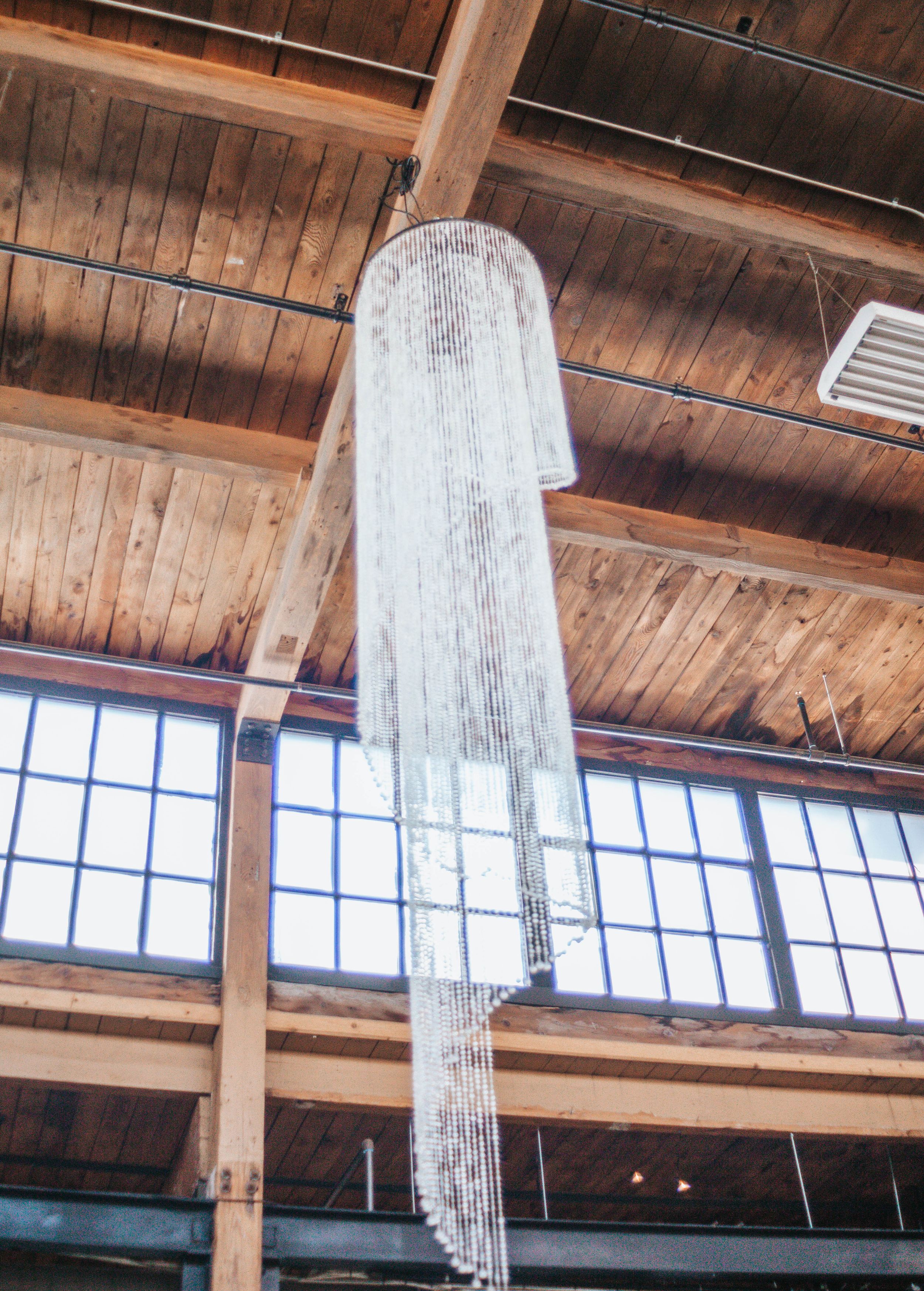 A modern crystal chandelier hangs from a rustic wooden ceiling with clerestory windows in an industrial-style space.