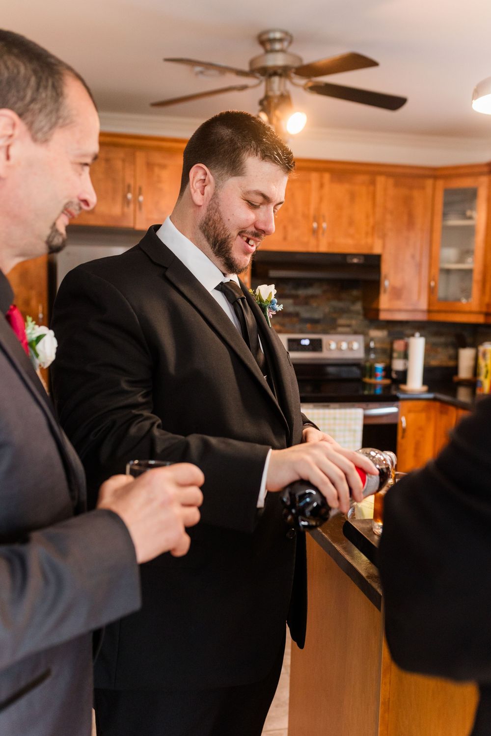 Groomsmen in black suits pour drinks into shot glasses in a wooden-paneled kitchen.