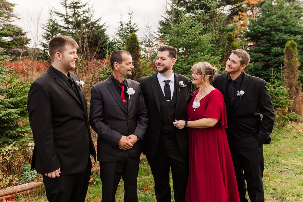 Wedding party members in black suits and a red dress interact joyfully outdoors among evergreen trees.