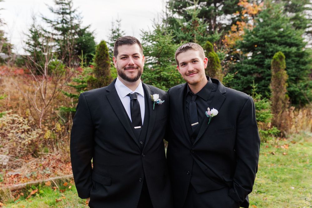 Two people in black suits stand together smiling outdoors with evergreen trees and autumn foliage in the background.