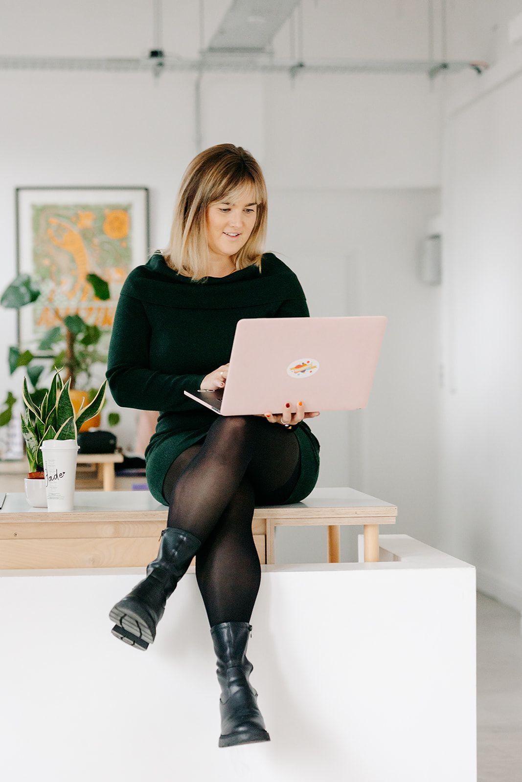 A person in a black sweater works on a pink laptop while sitting on a white desk in a bright modern office space.