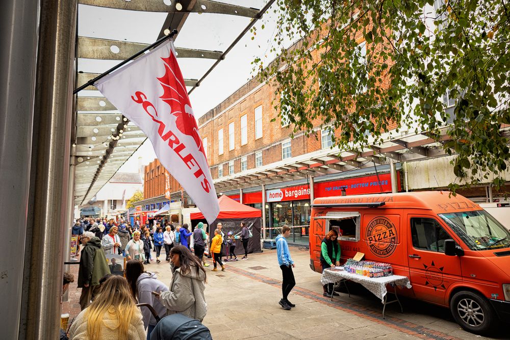 Red flags wave in front of brick buildings along a tree-lined street during a community event.
