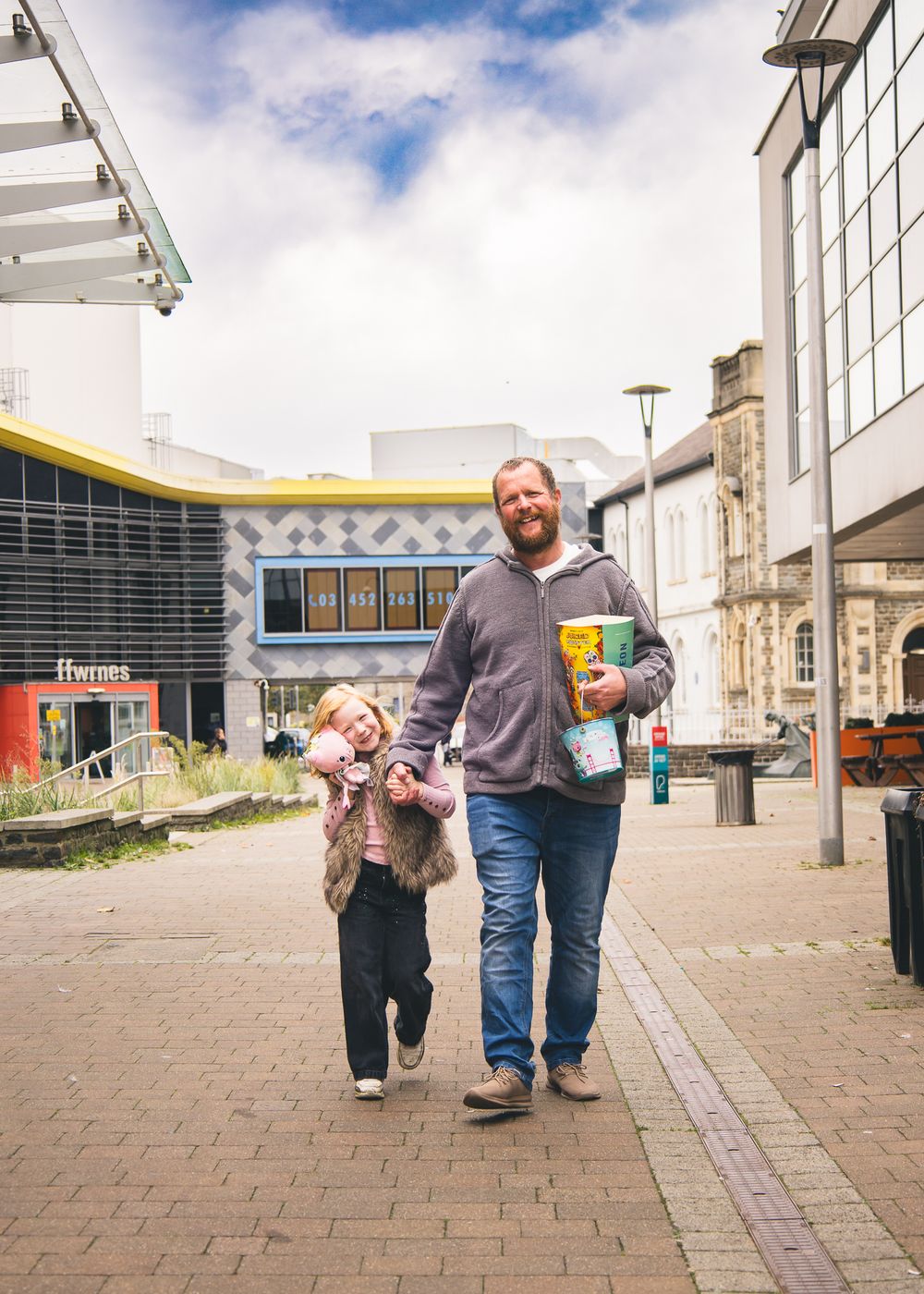 A parent and child walk together on a brick paved plaza outside modern buildings on a sunny day.