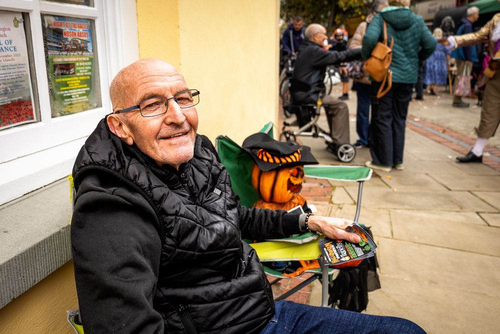 A person smiles while sitting in a wheelchair and wearing a black jacket at an outdoor event.