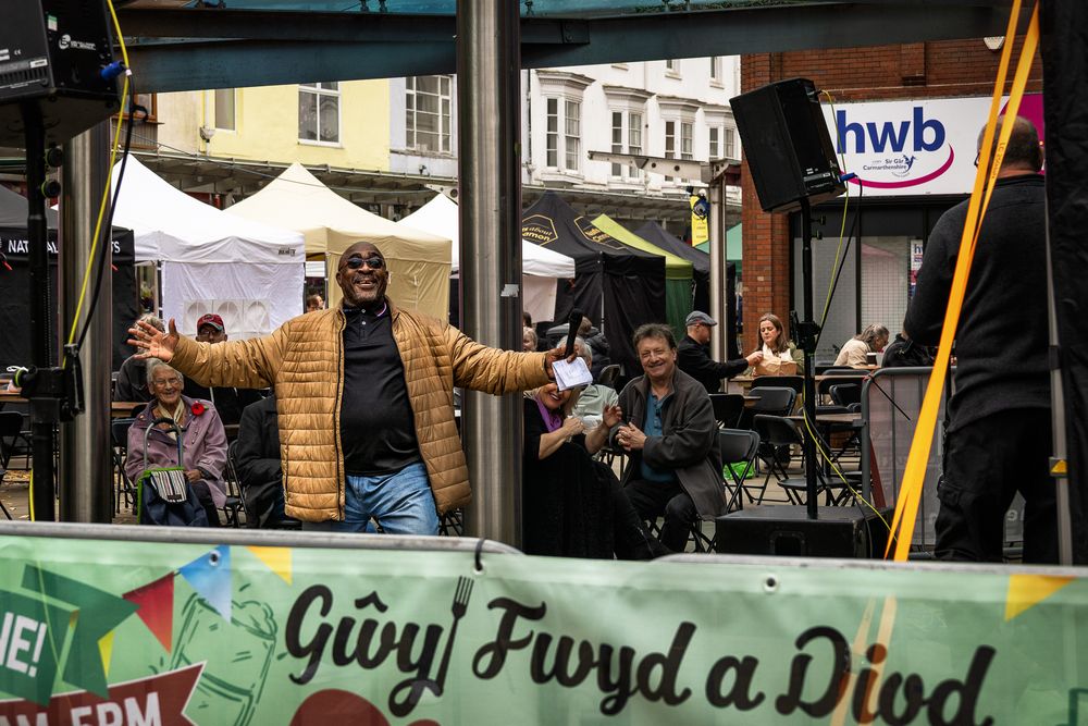 People browse outdoor market stalls with colorful displays and signs in what appears to be a Welsh marketplace.