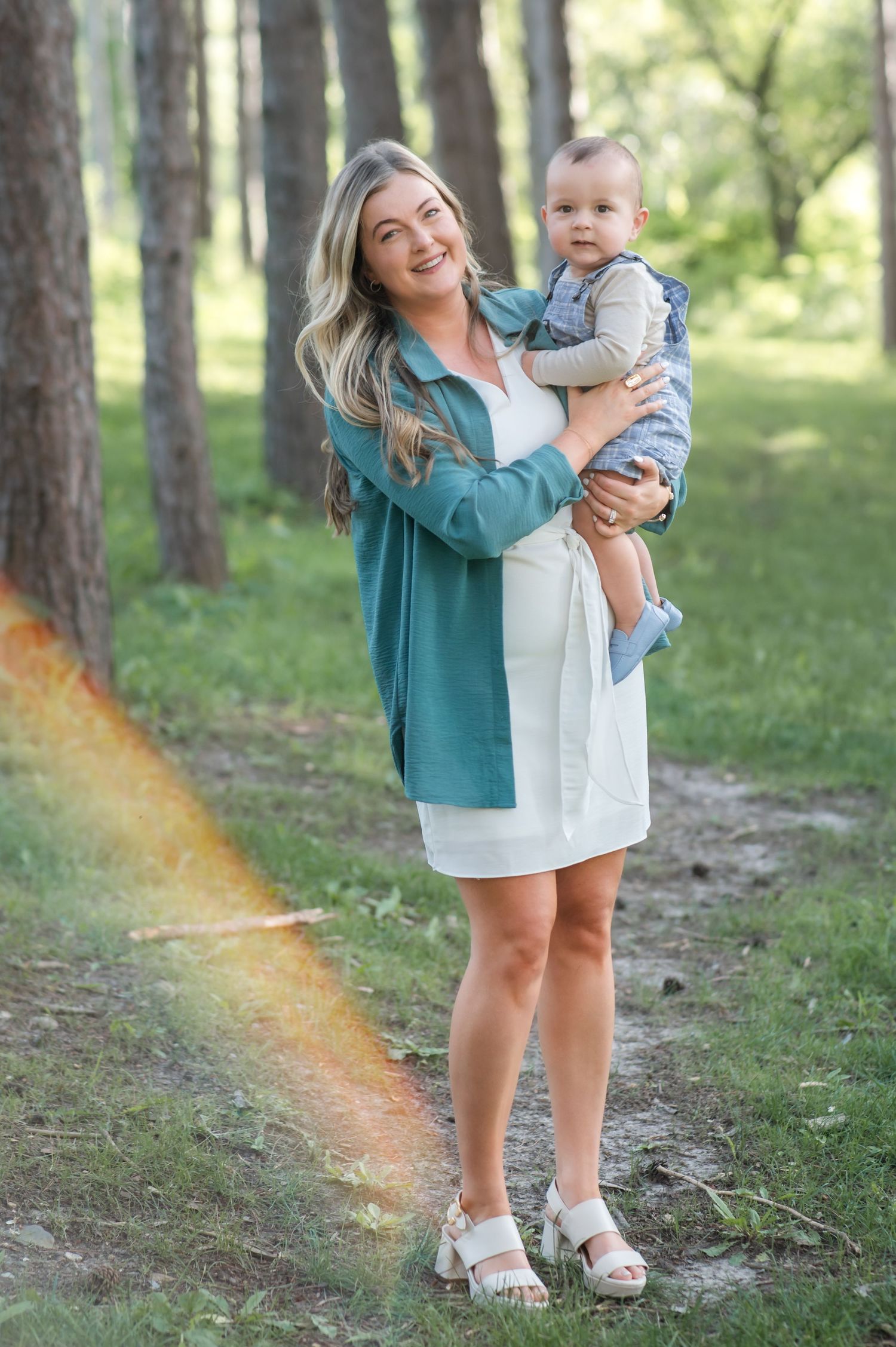 A woman in a teal cardigan plays with a baby outdoors in a sunlit forest setting.