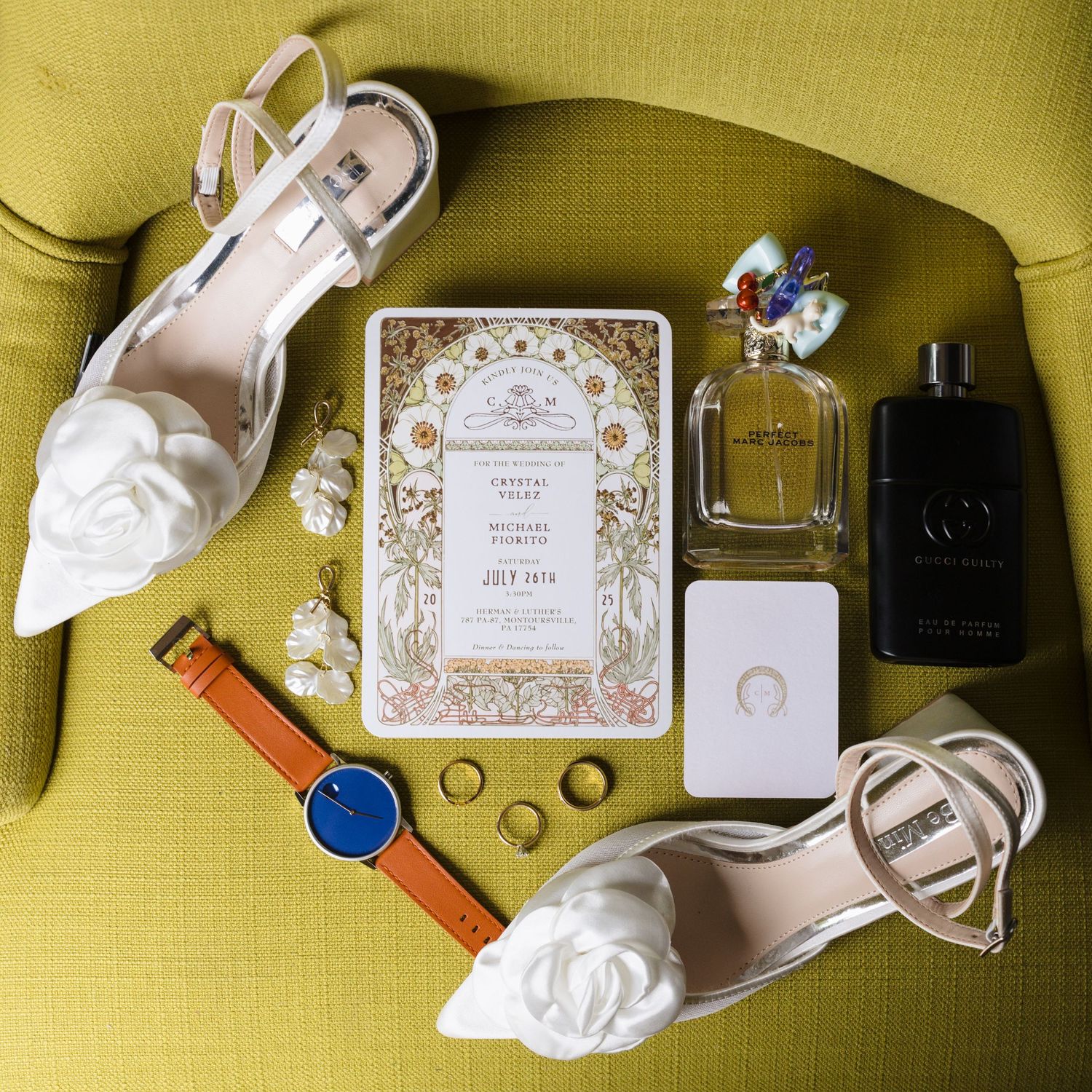Wedding details flatlay with white shoes, perfume bottles, invitation card, rings and orange watch on yellow background.