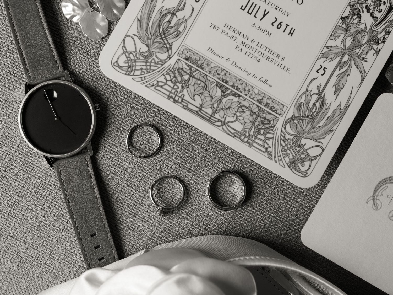 Black and white detail shot of wedding rings, invitation and watch on textured surface.