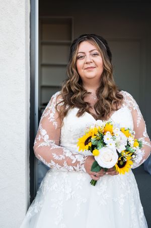 A bride in a white lace dress with sheer sleeves holds a vibrant sunflower bouquet against a light background.