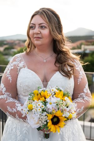 Bride in lace wedding dress holds sunflower and white flower bouquet during outdoor ceremony.