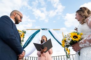 Outdoor wedding ceremony with participants reading vows against blue sky and mountain landscape.