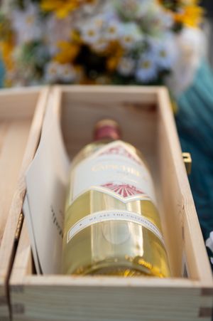 Wooden box displays wine bottles with decorative labels during outdoor wedding celebration.