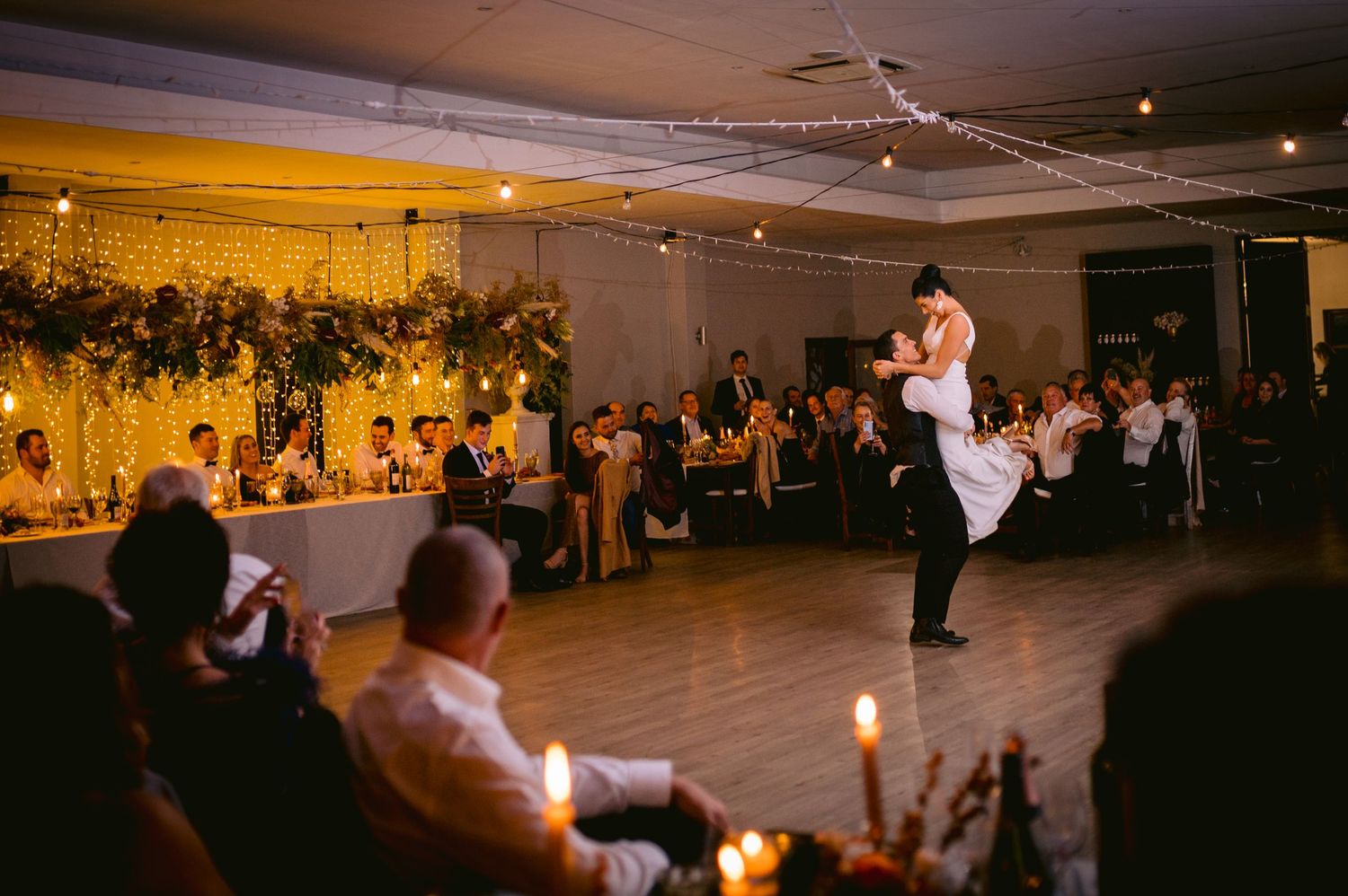 Wedding guests dance together in a dimly lit reception hall during an evening celebration.