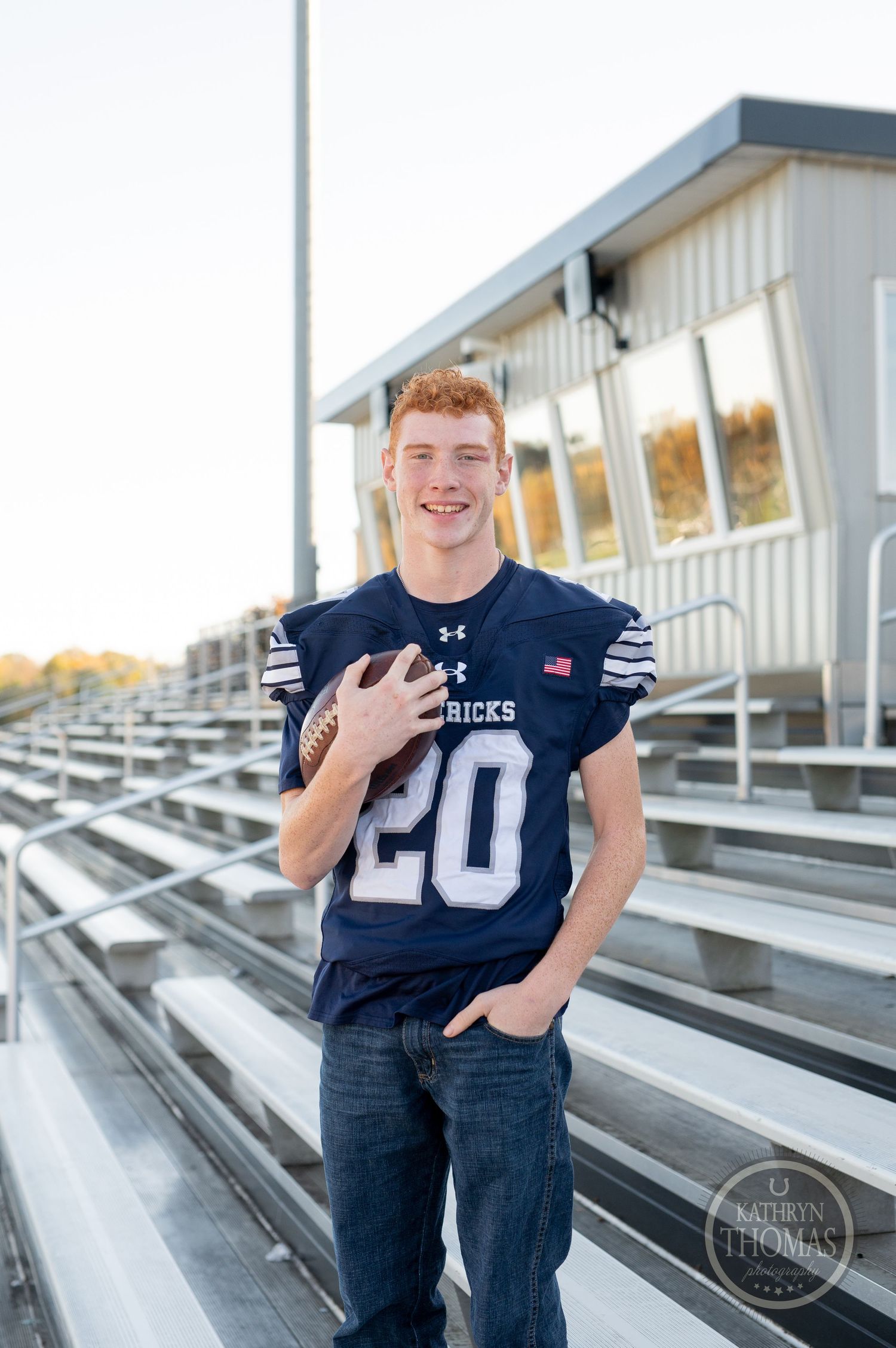 High school athlete poses in navy football jersey number 20 on stadium bleachers.