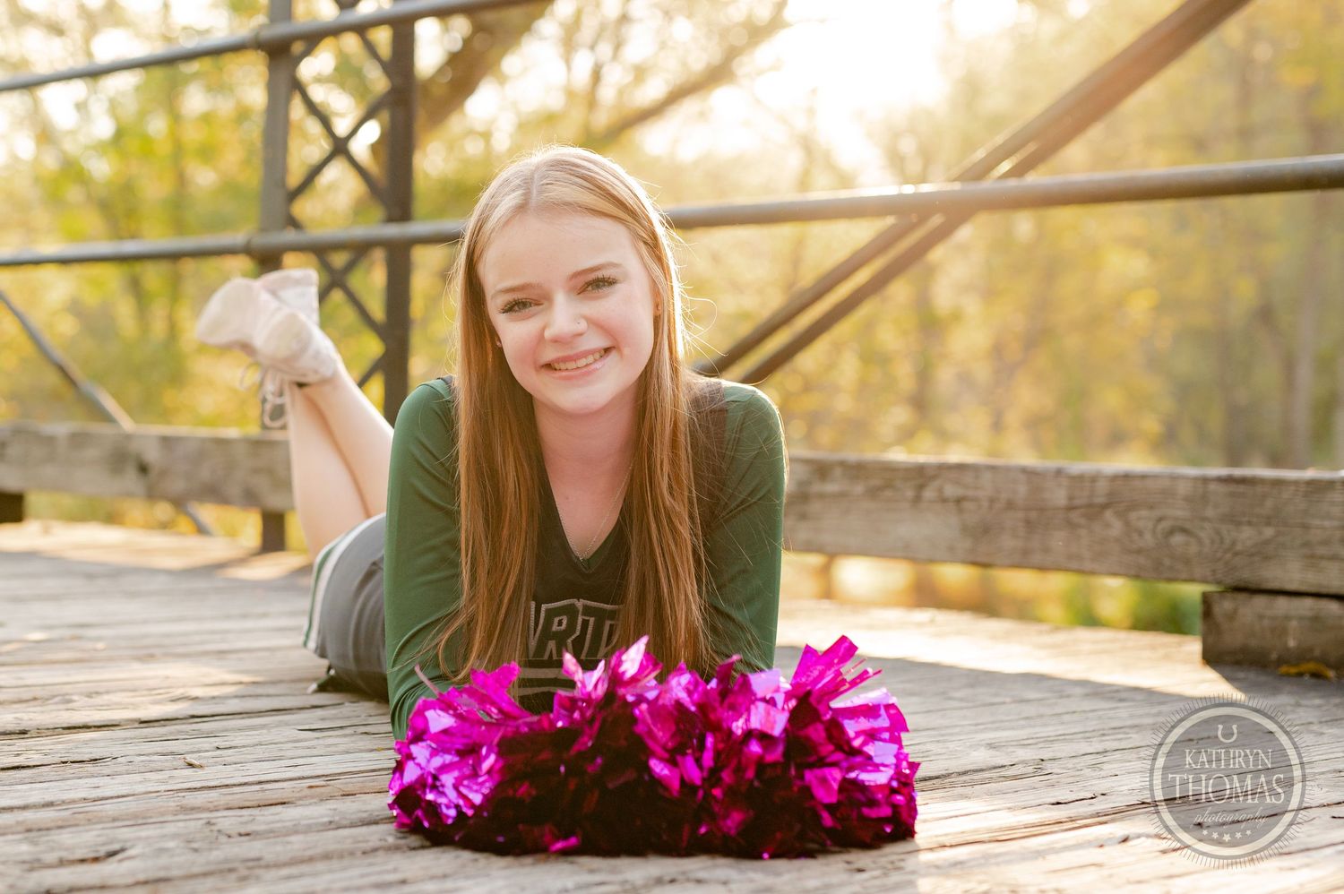 Cheerleader in green uniform lying on wooden bridge with bright pink pom poms at sunset.