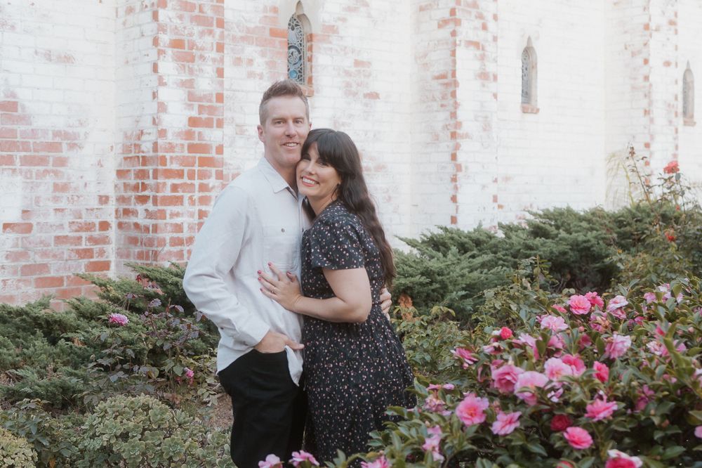 Romantic couple portrait against rustic brick wall with blooming pink flowers.