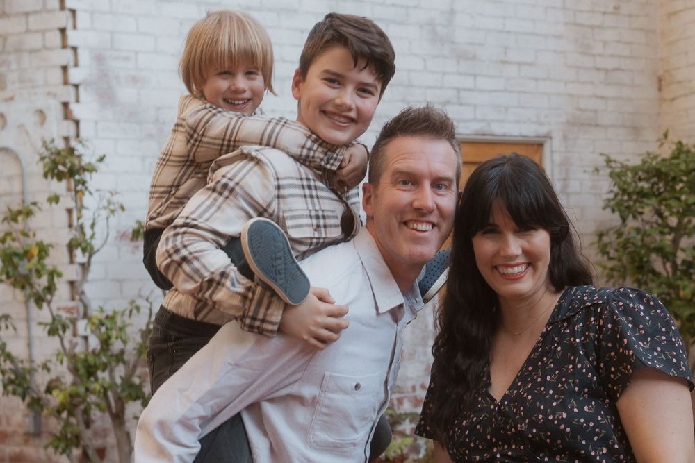 Family photo session against white brick wall with decorative elements and greenery in background.