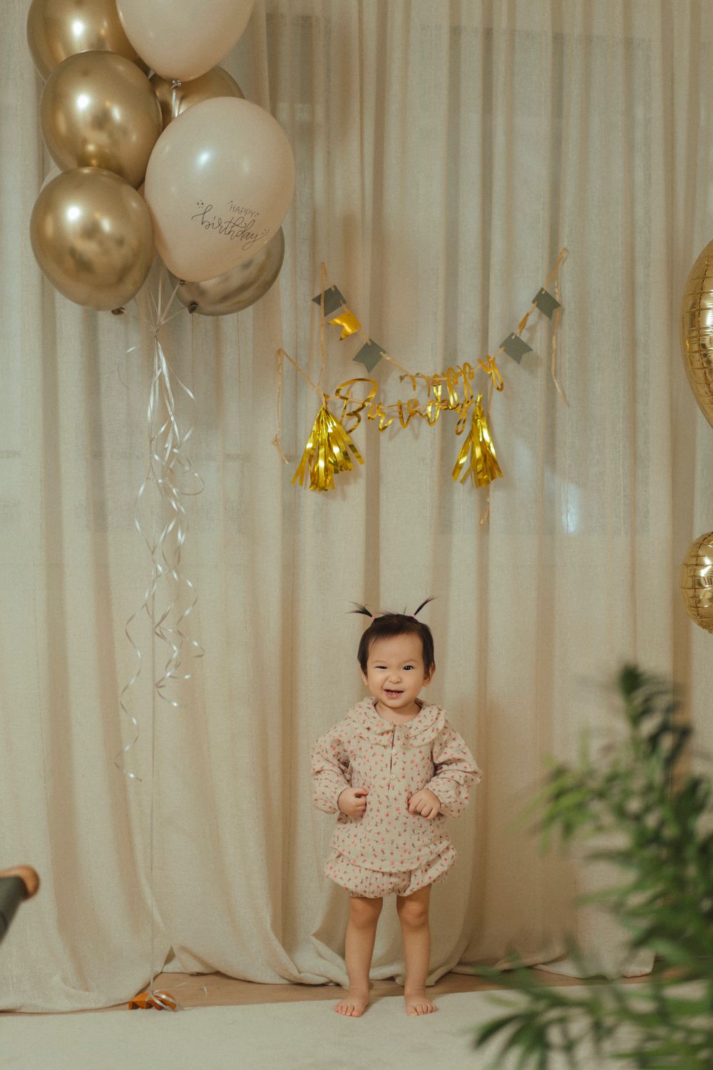 Elegant party setup with gold and white balloons, tassel garlands, and a beige curtain backdrop for a special celebration.