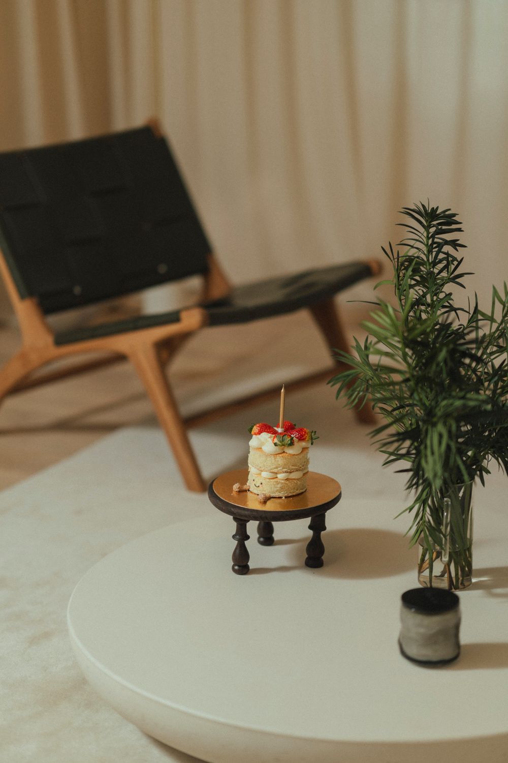 A small cake on a wooden pedestal sits beside a potted plant on a white round table with a lounge chair in the background.