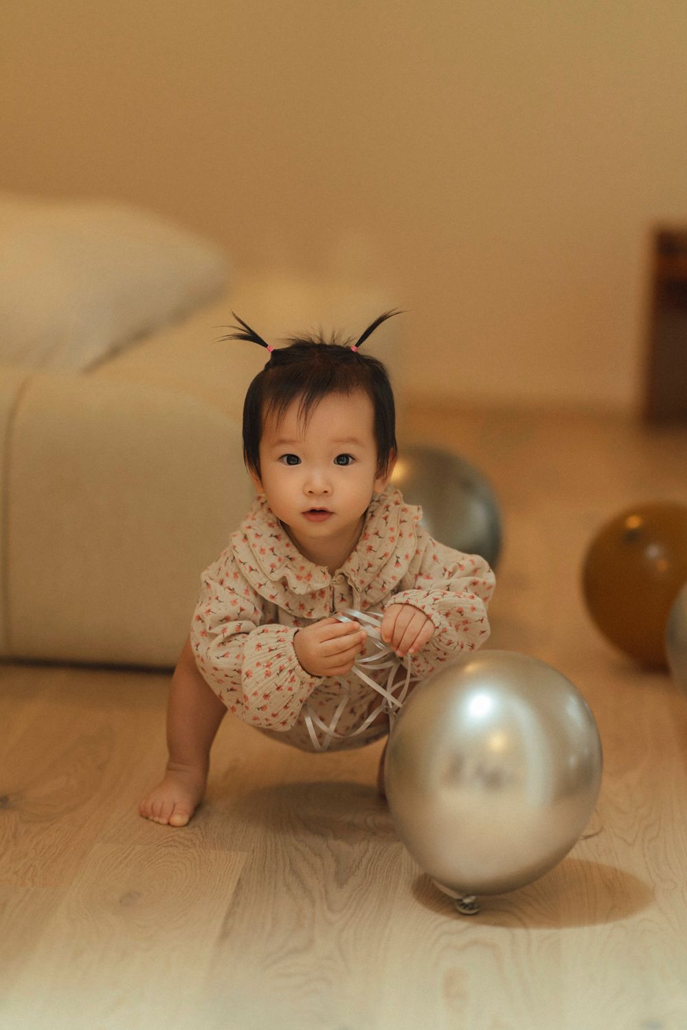 Small child in floral outfit crawls on wooden floor near silver balloons.