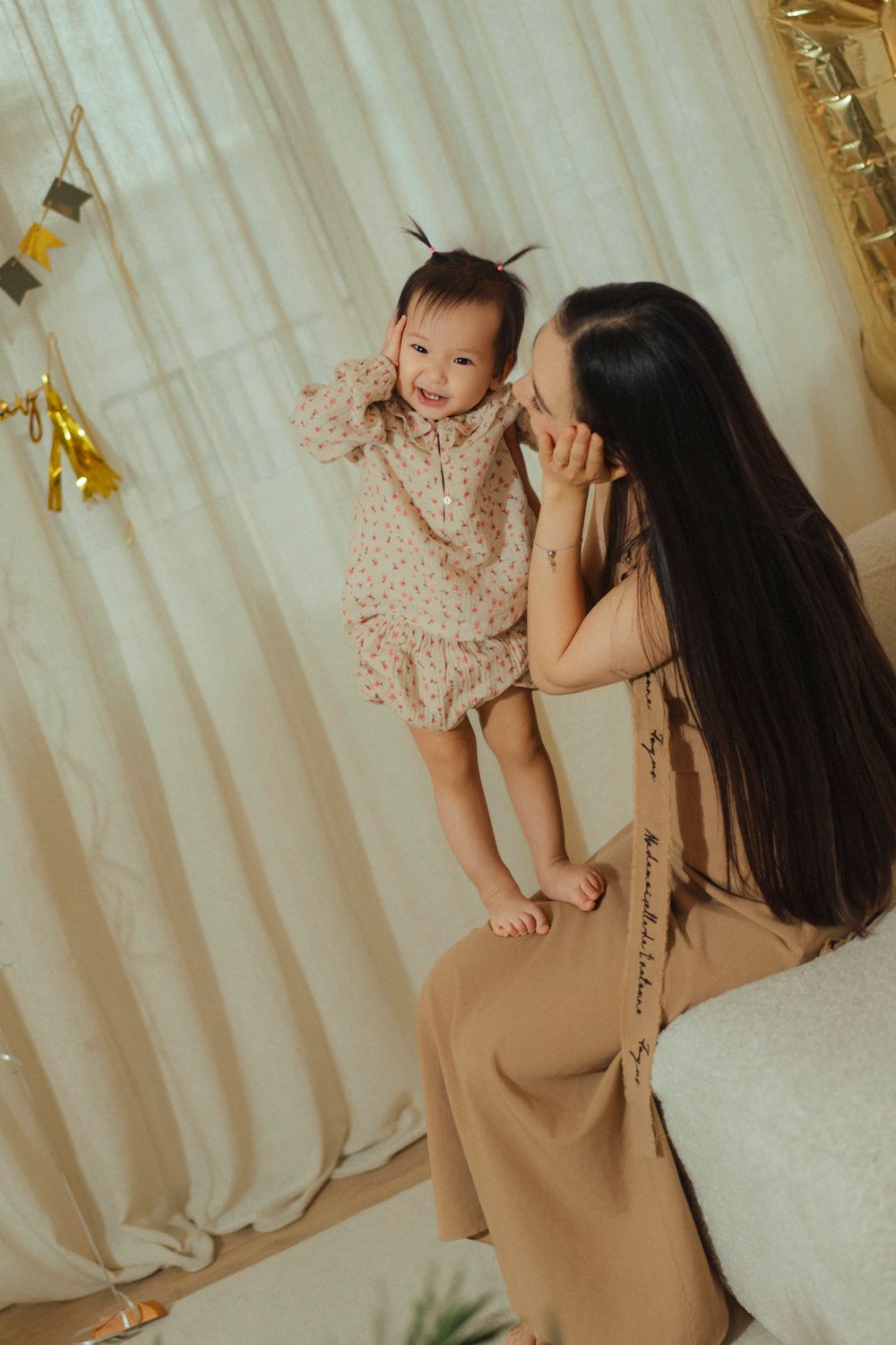A tender moment between parent and baby in polka dot dress against light curtains with gold party decorations.