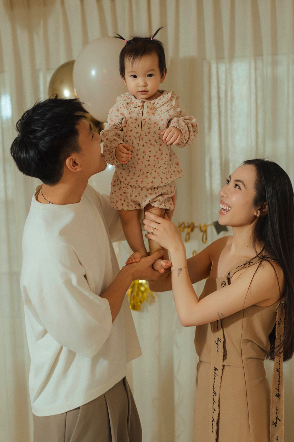 Family shares joyful moment lifting baby in polka dot outfit with balloon and curtain backdrop.