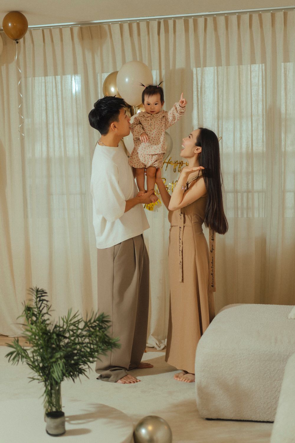 A family shares a joyful moment together in a minimalist room decorated with balloons and potted plants.