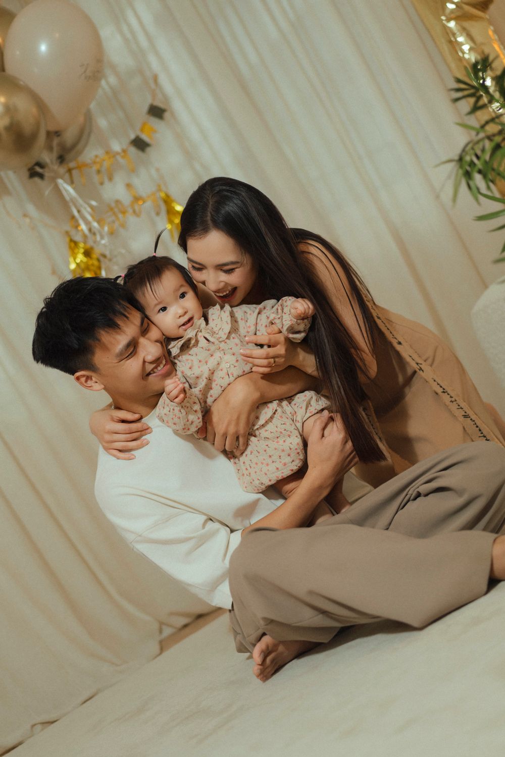 A family shares a tender moment together on a bed with soft white draping and balloons in the background.
