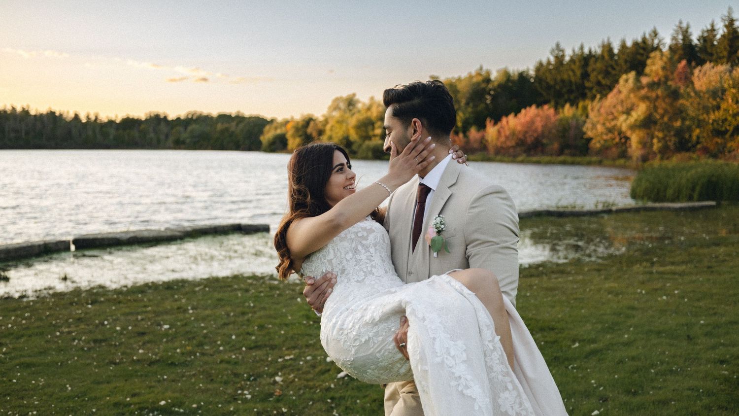 Groom carries bride in white dress by a lake with autumn trees at sunset during outdoor wedding celebration.