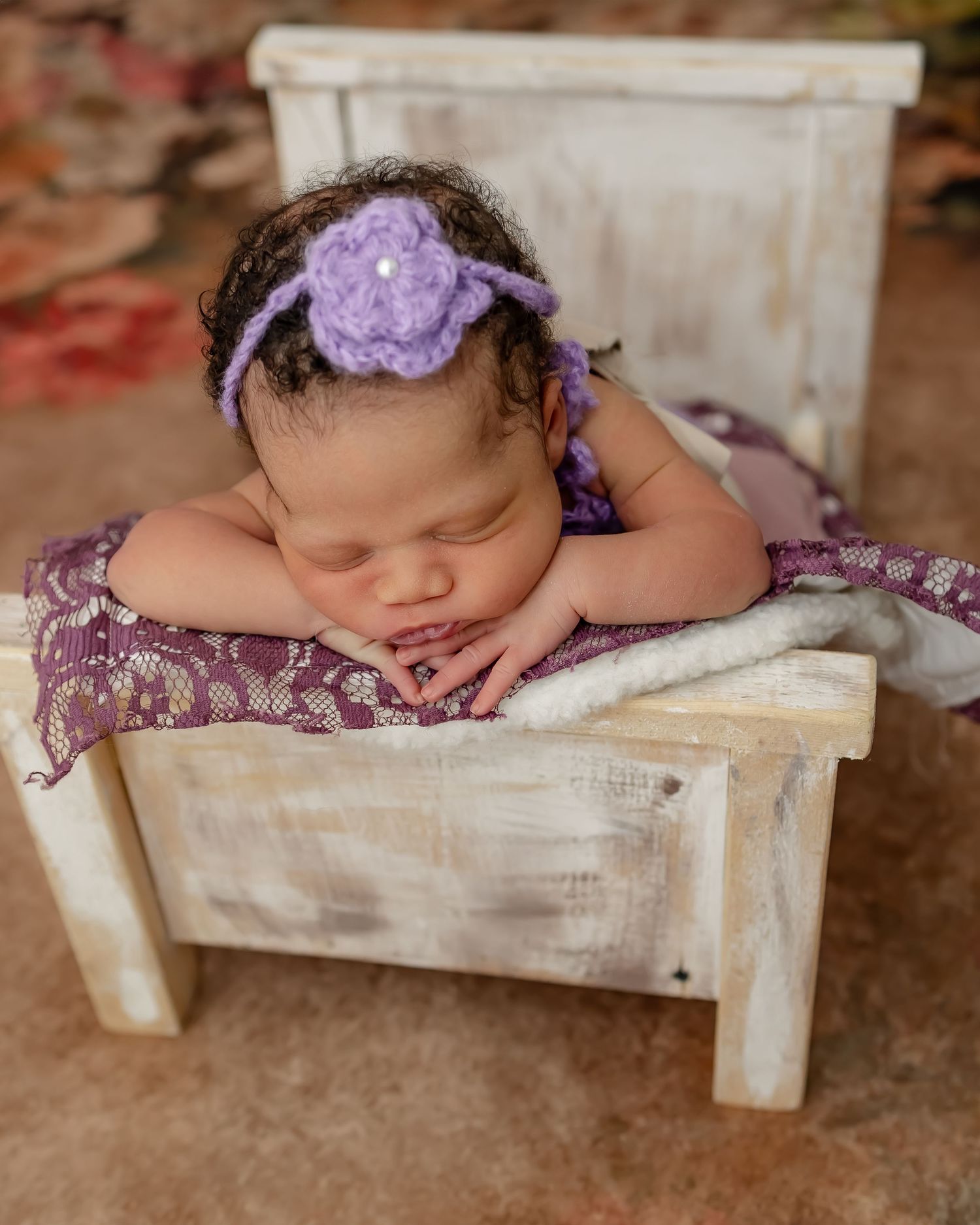 Sleeping baby with purple flower headband lying on white wooden bench.