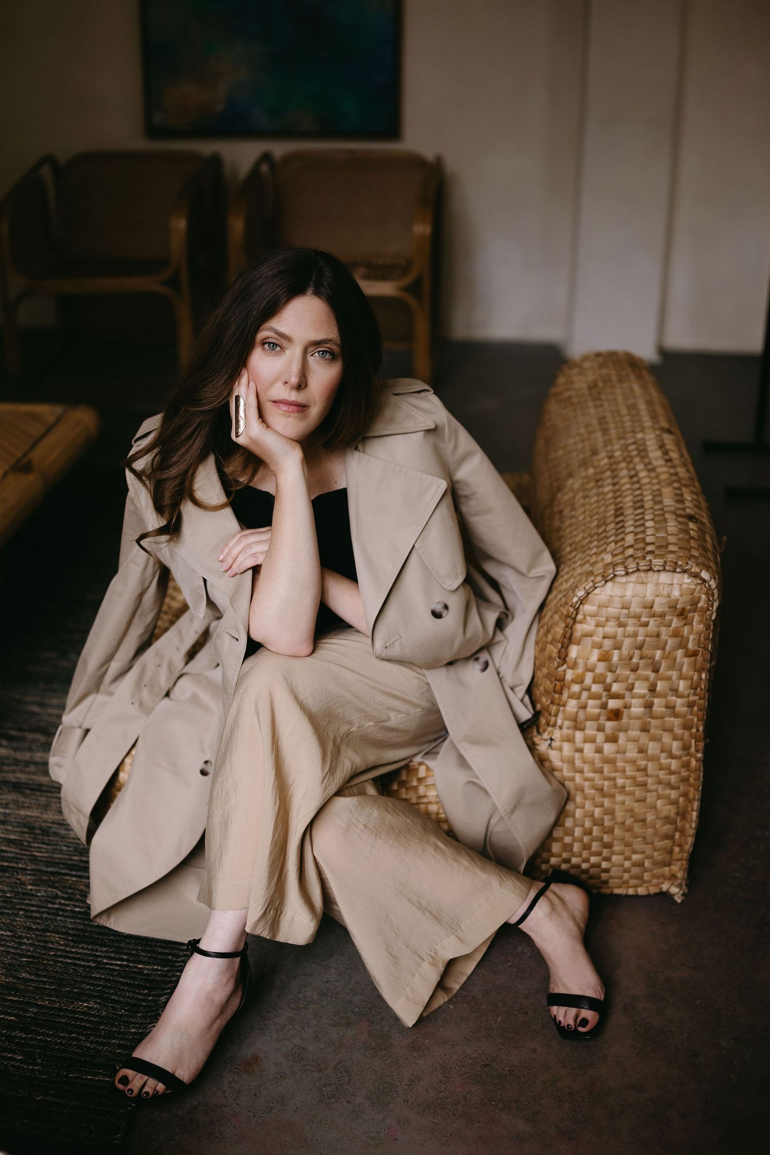 Fashionable beige trench coat styled with sandals on woven furniture in modern interior.