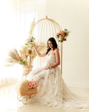 Ethereal maternity photoshoot featuring decorative birdcage, flowers, and flowing white lace gown.