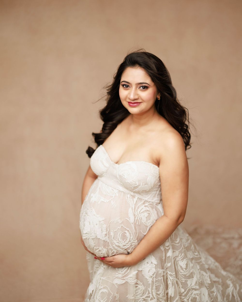 A maternity portrait featuring a white lace gown against a neutral brown backdrop.