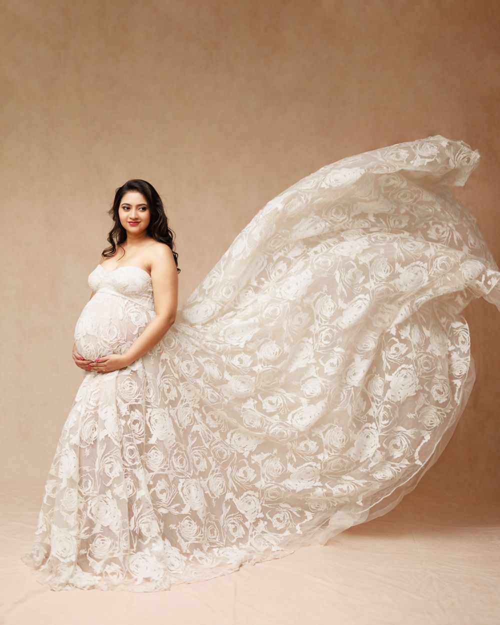 Beautiful lace maternity gown with flowing fabric shown against beige backdrop in elegant studio portrait.