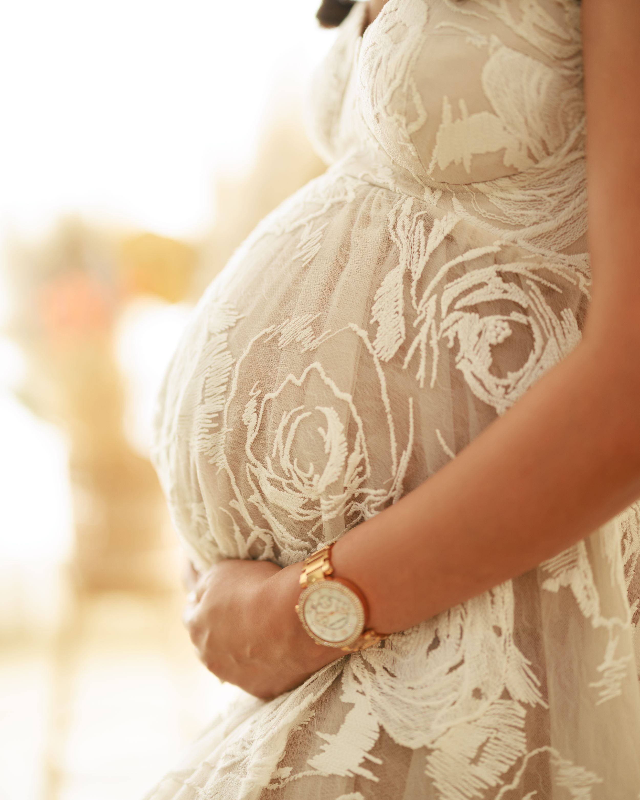 Close-up detail of textured rose pattern on white maternity gown with gold watch accessory.