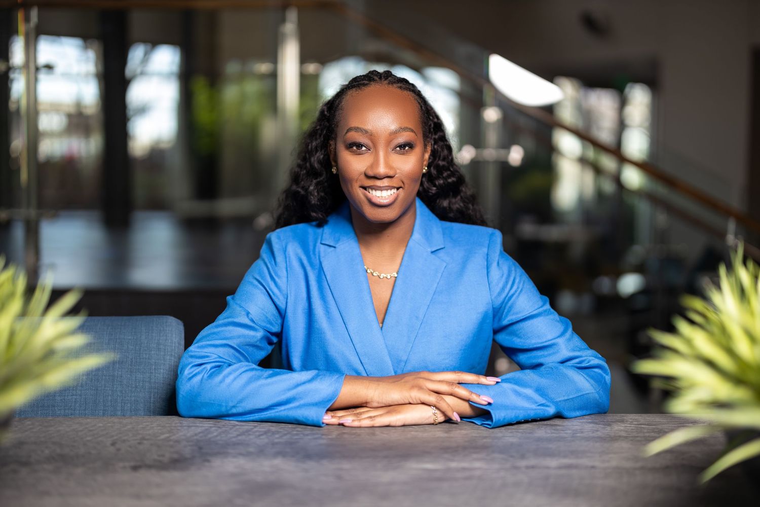 Professional in bright blue blazer seated at modern office desk with plants in background.