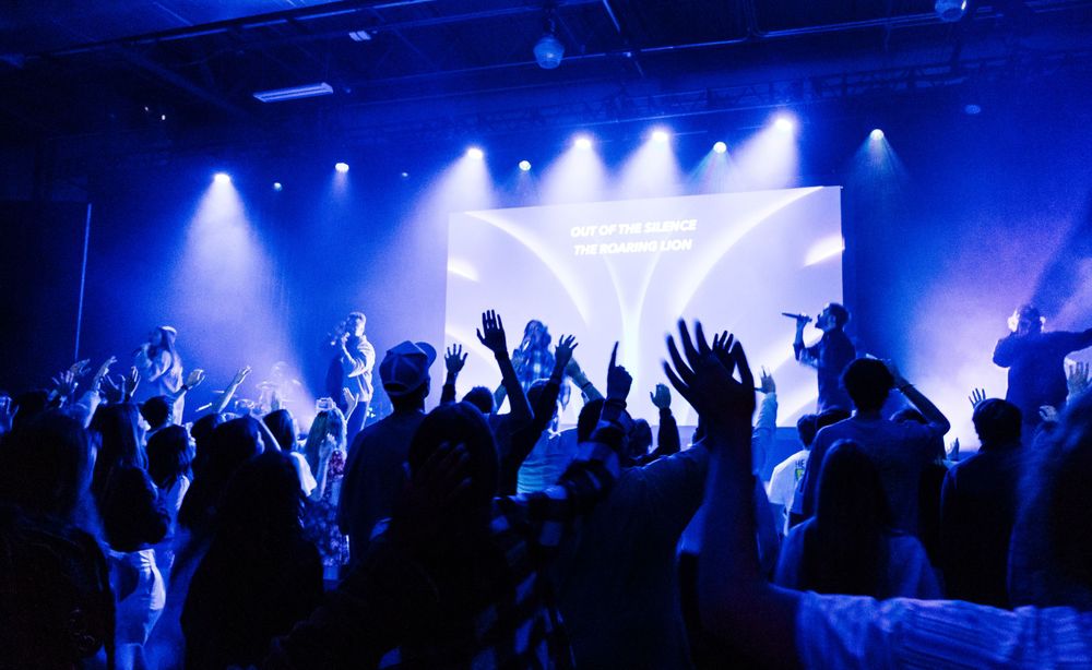 Concert crowd with raised hands in blue lighting at indoor music venue.