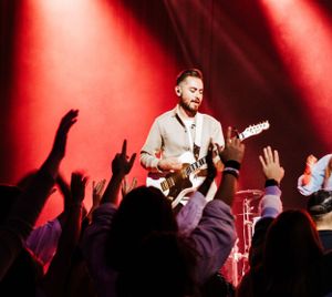 Rock musician playing electric guitar on stage with red spotlights and crowd raising hands.
