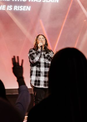 A worship leader performs on stage with red lighting and projected lyrics overhead.
