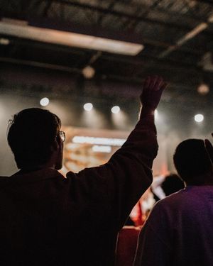 Silhouetted concert attendees raising hands during worship service in dim lighting.