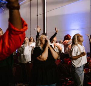 A group of energetic worshippers raising their hands in a concert setting with blue lighting.