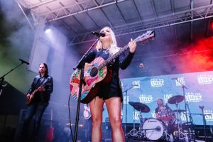 Female musician performing with acoustic guitar on outdoor stage with fog and red lighting effects.