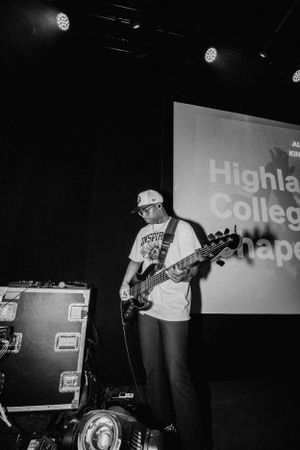 Bass guitarist performing on stage with Highland College projection in background.