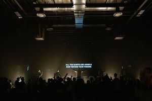 A silhouetted crowd at a concert with raised hands and lyrics displayed on screen.