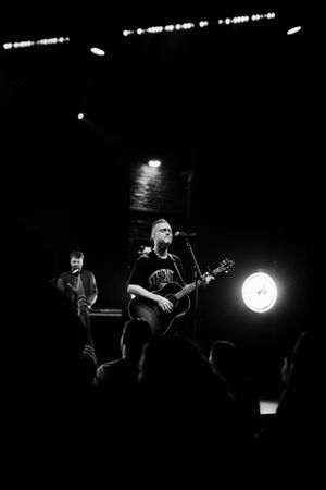Black and white photo of acoustic guitar performance on dimly lit concert stage.