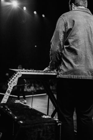 Black and white view of a musician's back showing a guitar headstock against stage lighting.