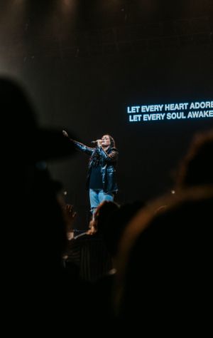 Concert performer silhouetted against blue text display during worship performance on dark stage.