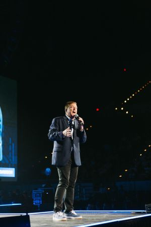 Solo performer speaking on illuminated stage during nighttime event with string lights in background.