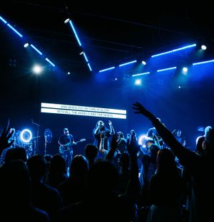 Concert crowd silhouettes against blue stage lighting with performers and text display visible.