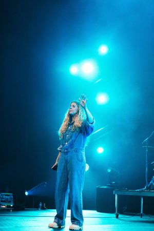 Performer in denim outfit singing under dramatic blue stage lighting at concert venue.